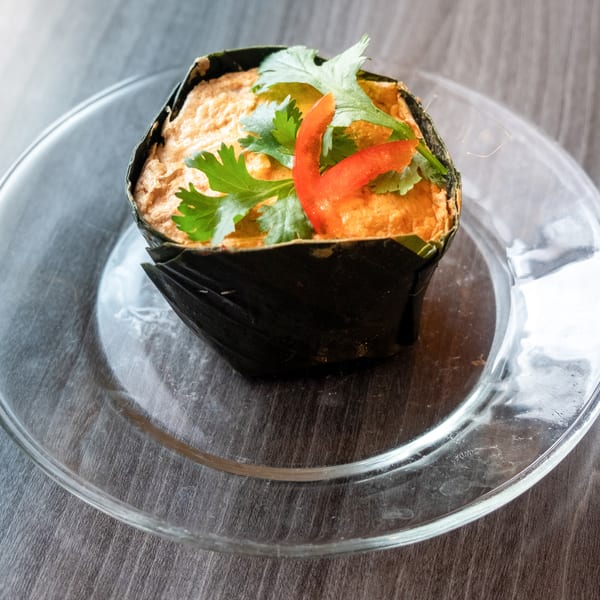 Steamed curry custard, wrapped in banana leaves, garnished with cilantro and red pepper on a glass plate.