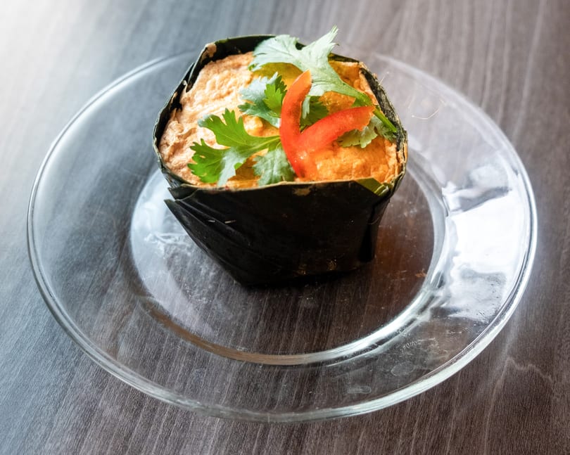Steamed curry custard, wrapped in banana leaves, garnished with cilantro and red pepper on a glass plate.