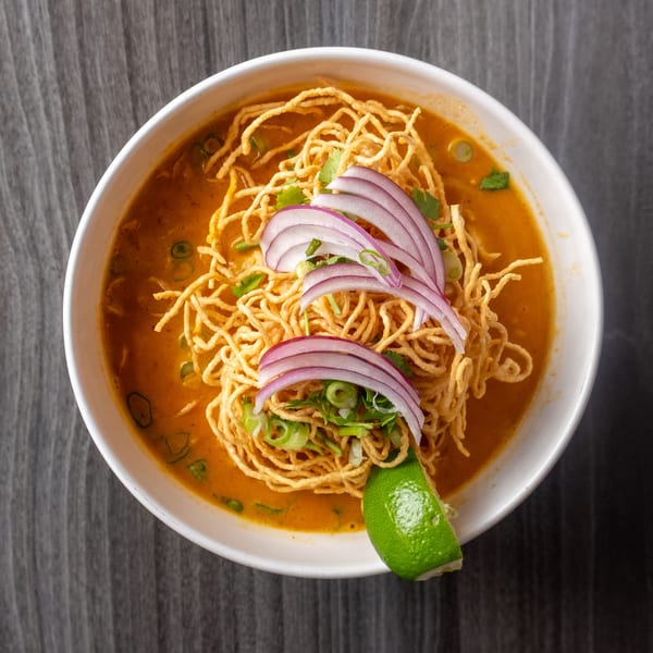 A bowl of Khao Soi with crispy noodles, red onions, green onions, and a lime wedge.