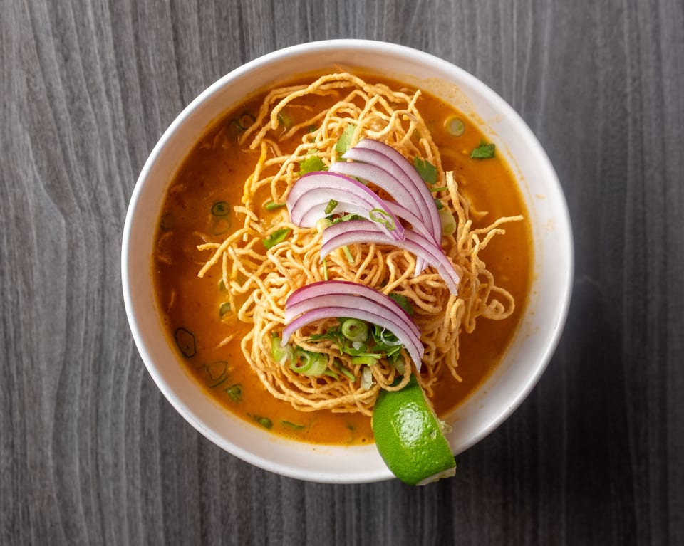A bowl of Khao Soi with crispy noodles, red onions, green onions, and a lime wedge.